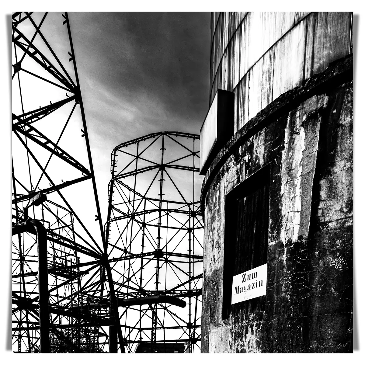 Schwarz-weiß-Fotografie einer verlassenen Industrieanlage mit Stahlgerüsten und einem alten Betonsilo. Im Vordergrund ein verwittertes Schild mit der Aufschrift „Zum Magazin“. Die Szenerie wirkt verlassen, rau und eindrucksvoll.