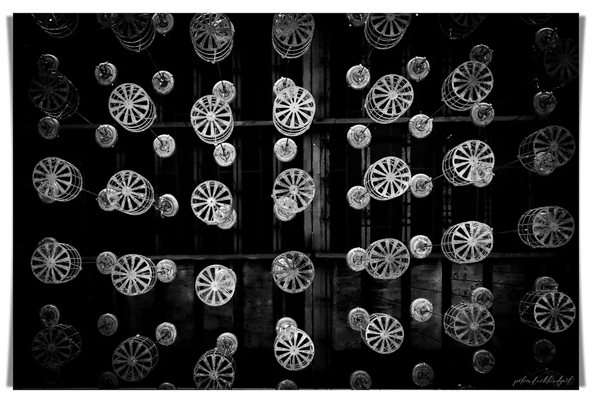 Leere Drahtkörbe in einer alten Zechen-Waschkau hängen an Seilzügen von der Decke. Schwarzweißaufnahme, symmetrisch und ruhig.