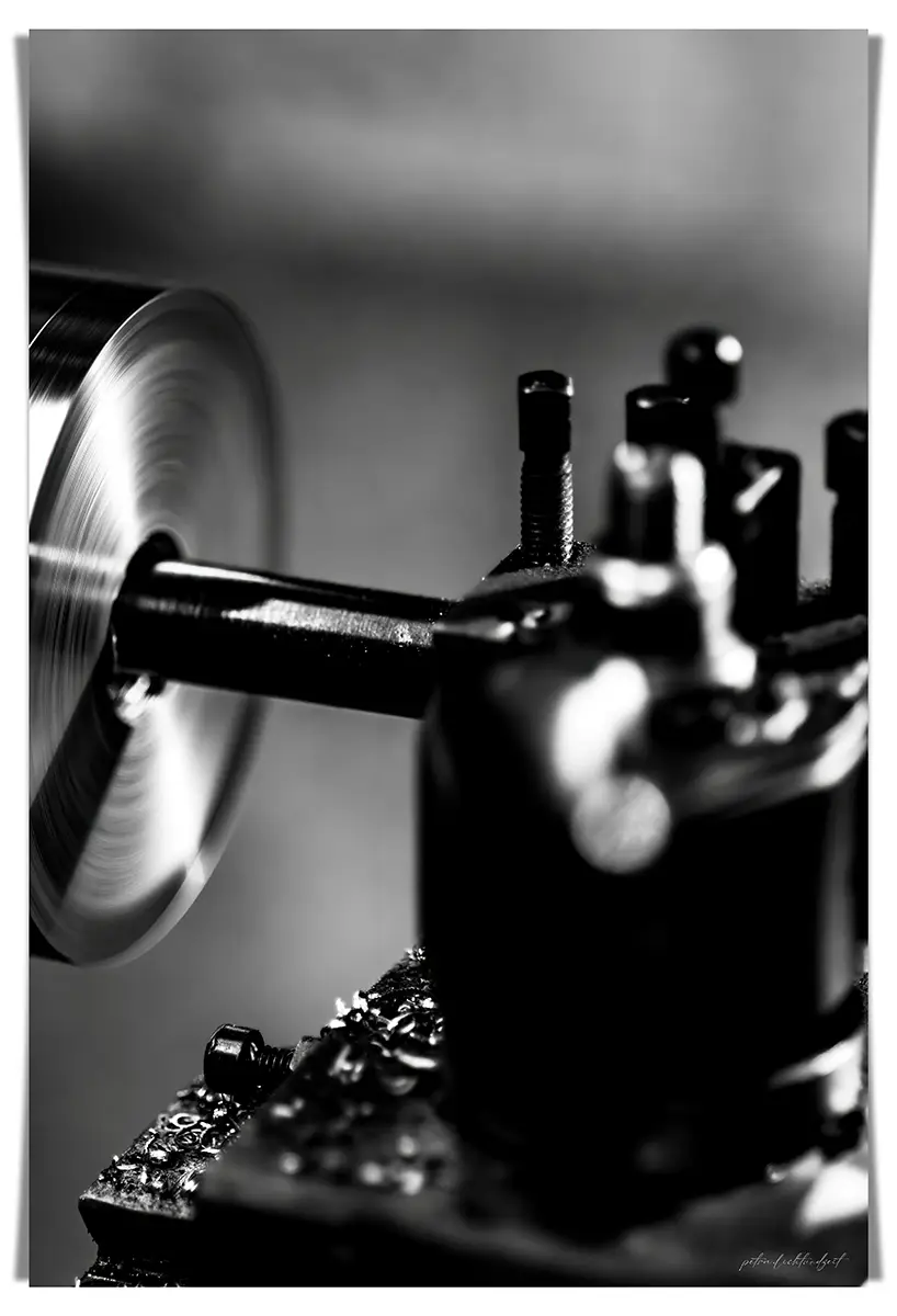 Nahaufnahme einer laufenden Drehmaschine in Schwarz-Weiß. Links ist eine rotierende Metallscheibe mit deutlich sichtbarer Bewegungsunschärfe zu sehen. Im Vordergrund befinden sich unscharfe Maschinenteile und Metallspäne. Der Hintergrund ist neutral und verschwommen.