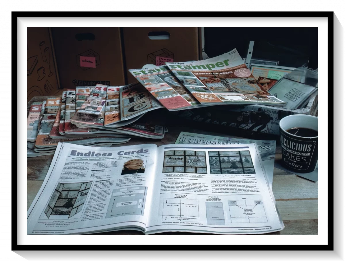 Ein Stapel internationaler Kreativzeitschriften mit einem aufgeschlagenen Artikel über Endloskarten. Im Hintergrund Koffer, Kisten und eine Tasse Kaffee.