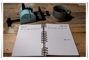 Der fertig gebundene KaBuJo 2026 liegt geöffnet auf einem Holztisch. Daneben eine Tasse Kaffee und das türkisfarbene Bindegerät. Die Seiten zeigen die Woche 10 und 11.