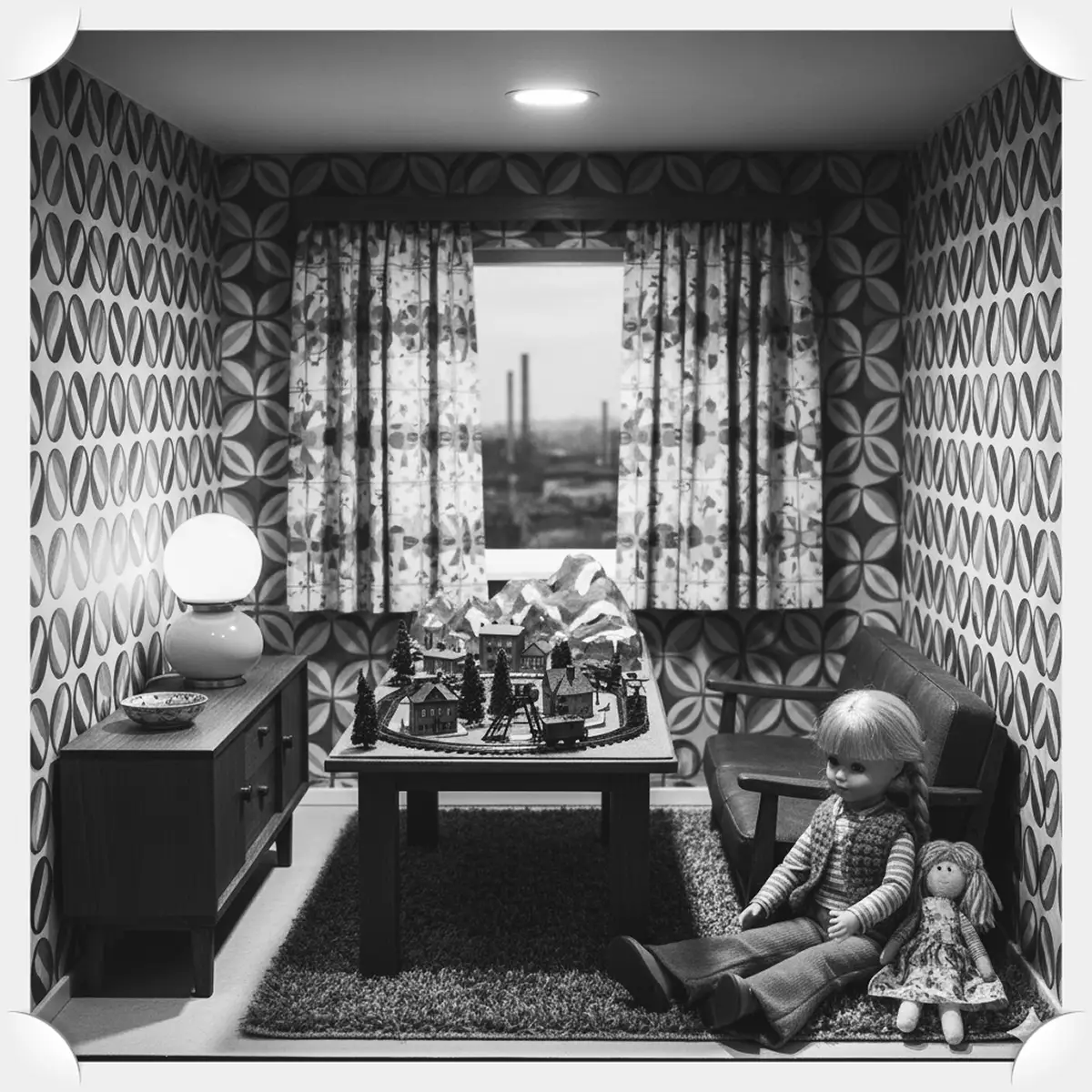 Puppenstube mit Modelleisenbahn im Stil der 1970er: Ein Mädchen mit Zöpfen sitzt vor einem Tisch, auf dem eine Modelllandschaft mit Bergen und Schienen aufgebaut ist. Im Hintergrund stehen ein Sideboard mit Lampe, ein Sofa und ein Fenster mit Blick auf Schornsteine im Ruhrgebiet. Die Einrichtung ist retro und detailreich.
