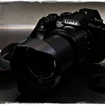 Panasonic FZ1000 II Superzoom-Bridgekamera mit Gegenlichtblende, frontal aufgenommen. Seitenansicht der Panasonic FZ1000 II Kamera mit Handgelenks-Trageriemen.