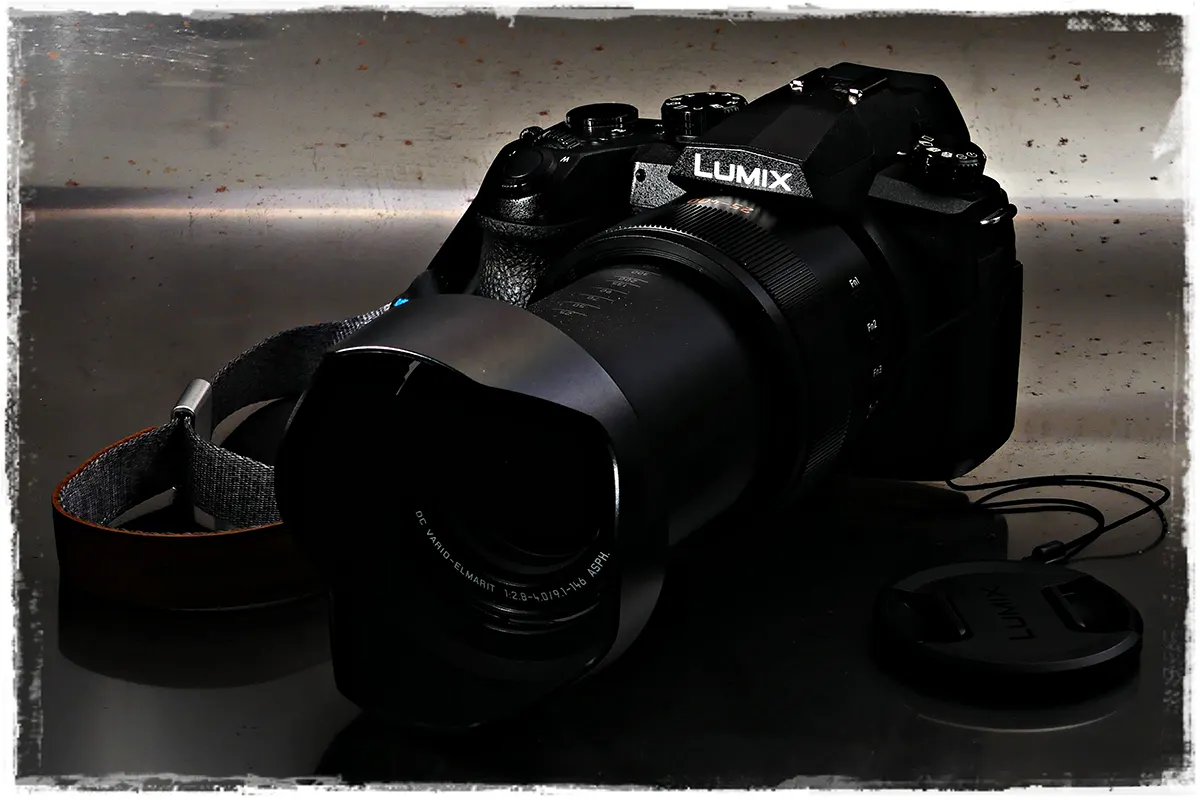 Panasonic FZ1000 II Superzoom-Bridgekamera mit Gegenlichtblende, frontal aufgenommen. Seitenansicht der Panasonic FZ1000 II Kamera mit Handgelenks-Trageriemen.
