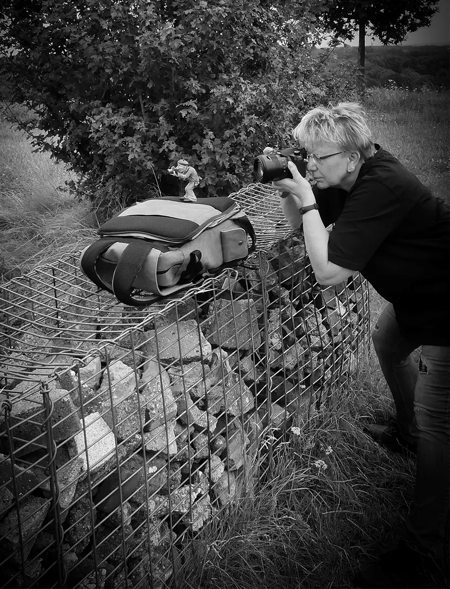 Petra beim Fotografieren einer Spielfigur im Freien, Kamera auf einem Stein-Gabionenstapel abgestützt, humorvolle Szene in Schwarz-Weiß.