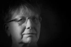 Schwarz-weiß Porträtaufnahme von Petra, die ernst, ruhig und direkt in die Kamera blickt – mit Brille, Kurzhaarfrisur und weichem Licht von links.