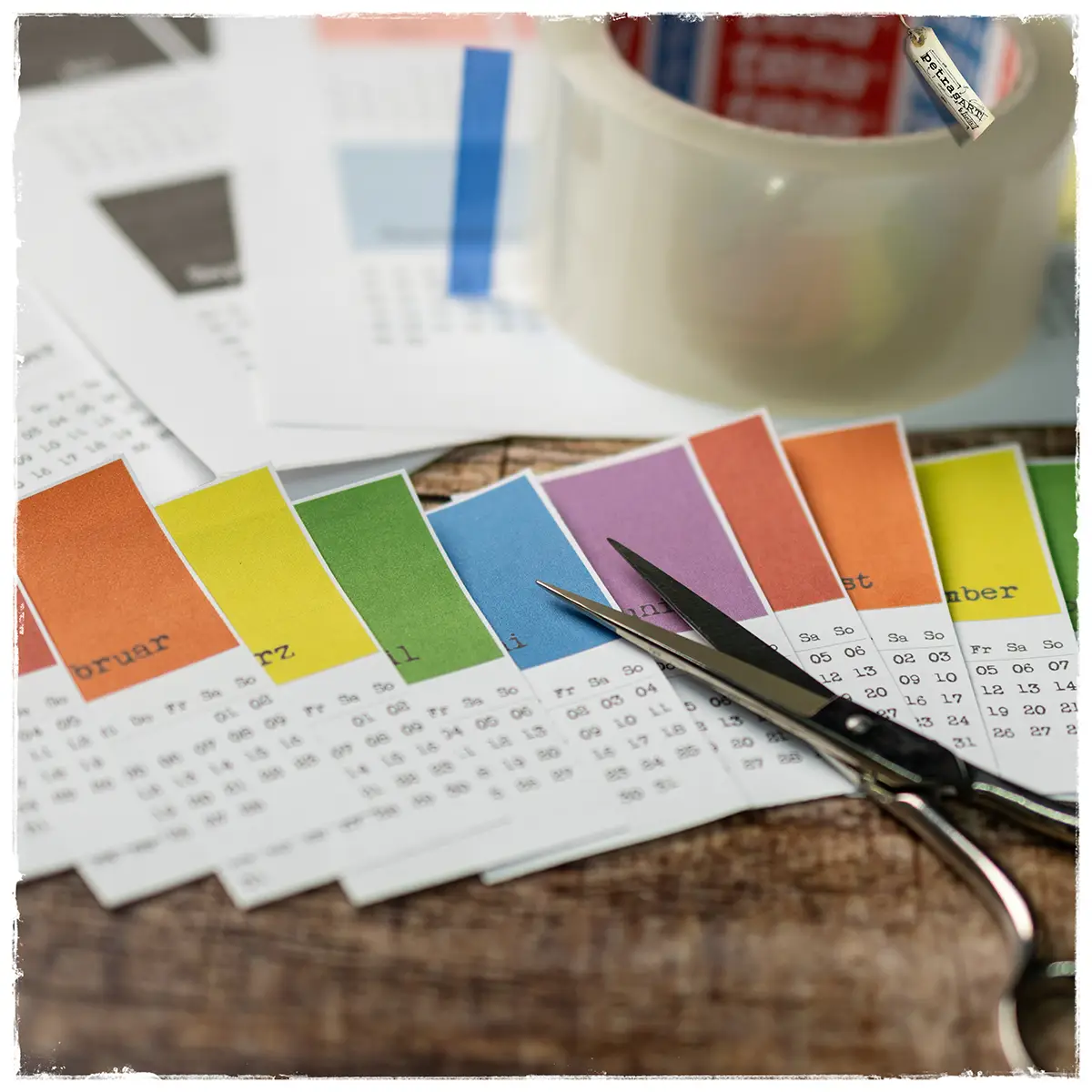 Farbstreifen-Minikalender ausgeschnitten und bereit zur Verarbeitung – mit Schere, Etikettenpapier und Klebeband.